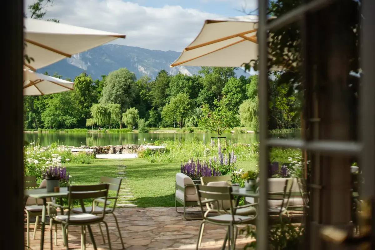 View from a cozy garden terrace with a seating area and sun umbrellas across a wildflower meadow and the castle pond toward the Salzburg Alps, in the Leopoldskron Palace Garden.