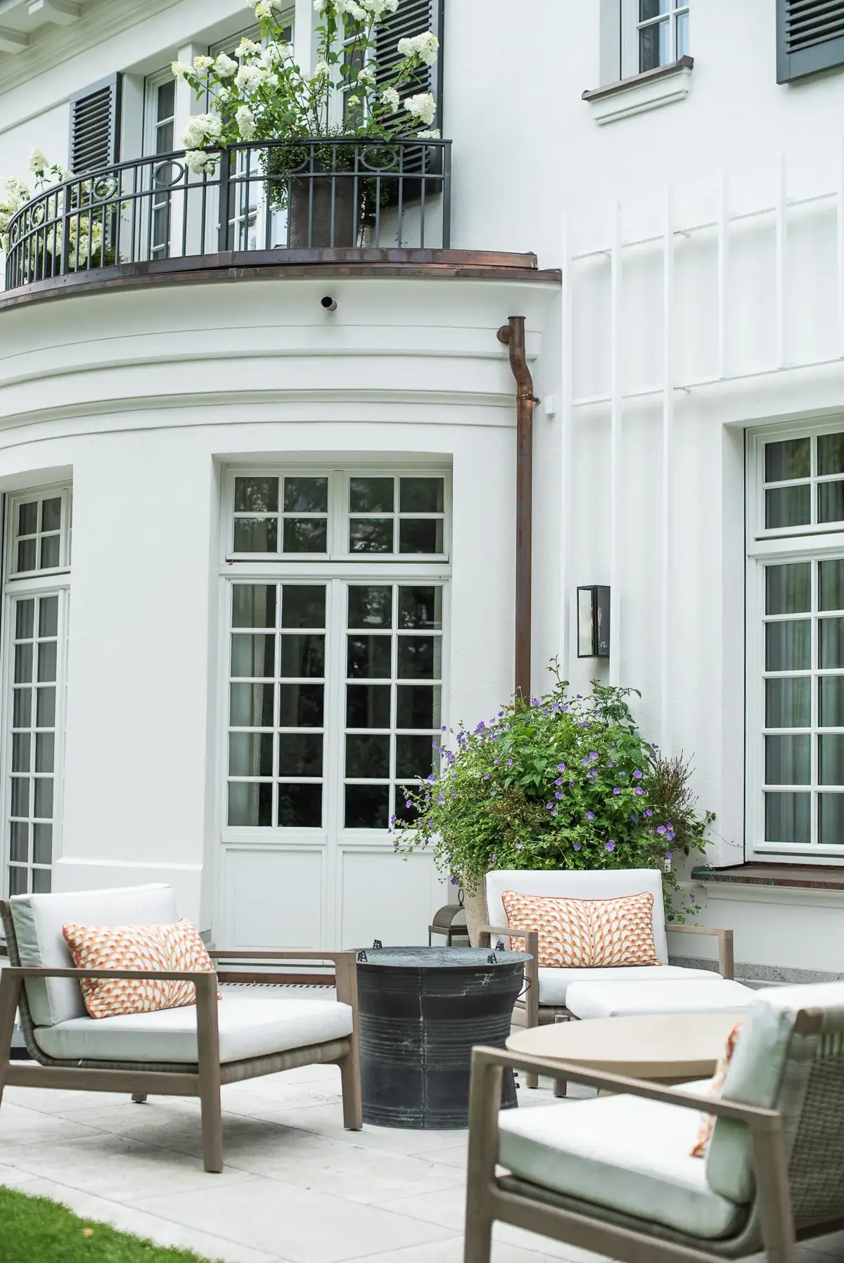 Gerundeter Fassadenerker mit schmiedeeisernem Balkon und weißen Hortensien, darunter eine Loungeterrasse mit gepolsterten Sitzmöbeln.