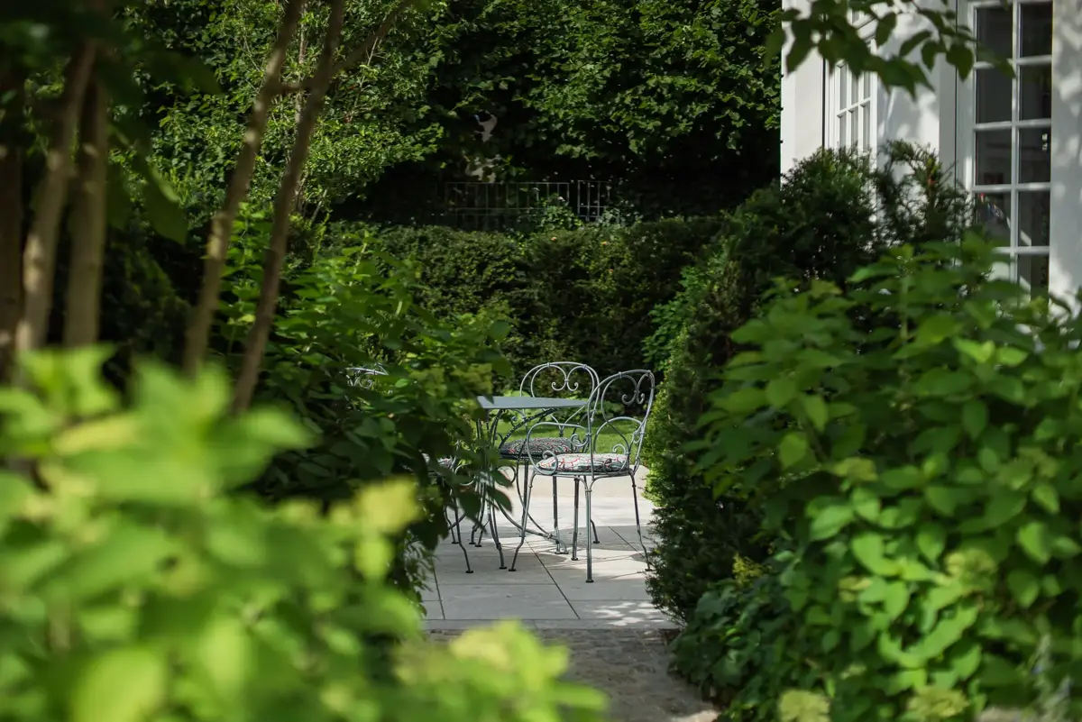 Blick durch dichte Hortensien- und Heckenpflanzung auf eine kleine Terrasse mit Bistromöbeln vor der weißen Fassade der Stadtvilla.
