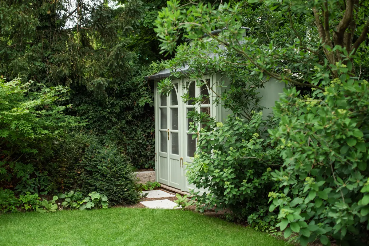Verstecktes Gartenhaus mit salbeigrünen Flügeltüren und goldenen Beschlägen, eingebettet in Japanischen Ahorn und Eibenhecken im Villengarten München.