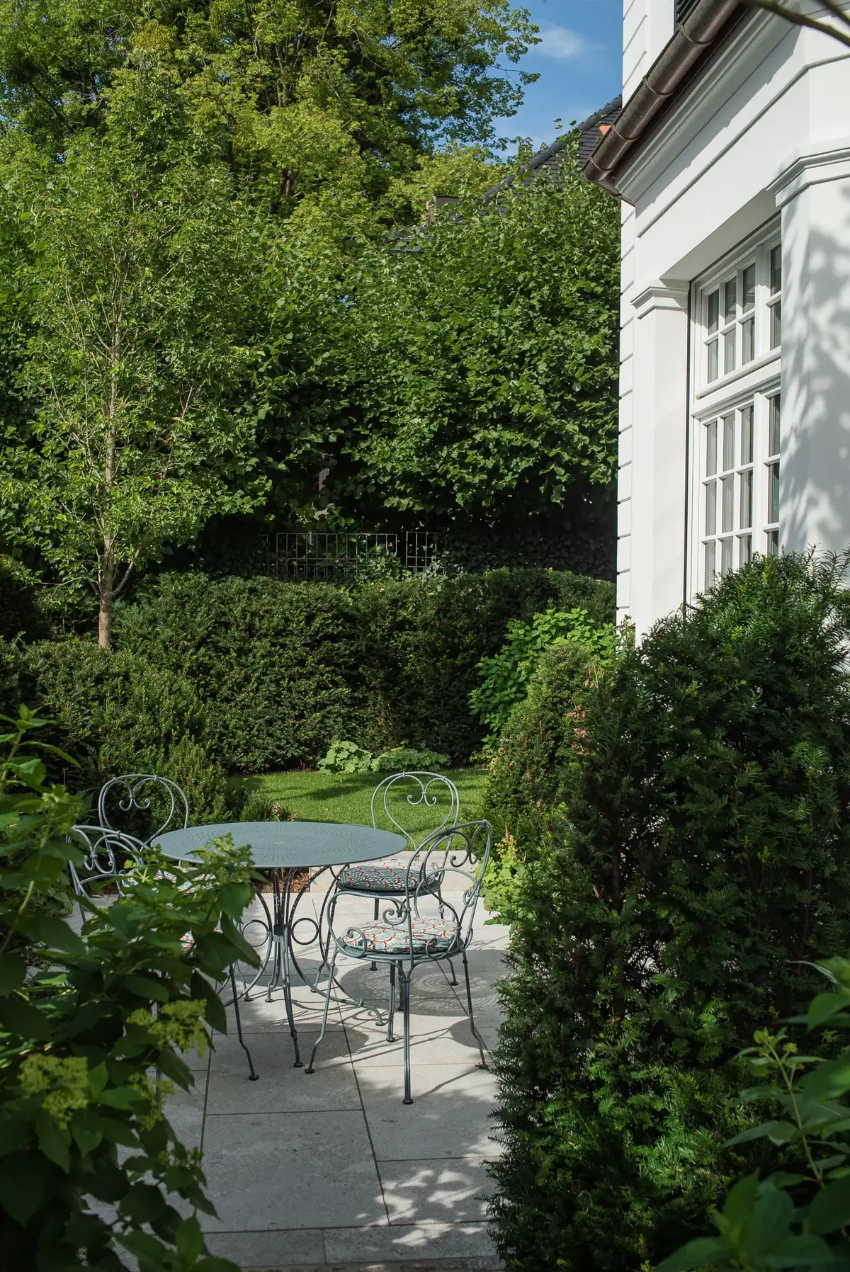 Kleiner, geschützter Sitzplatz mit schmiedeeisernem Bistro-Tisch zwischen hohen Eibensäulen und dichter Hainbuchenhecke im klassischen Villengarten München-Bogenhausen.
