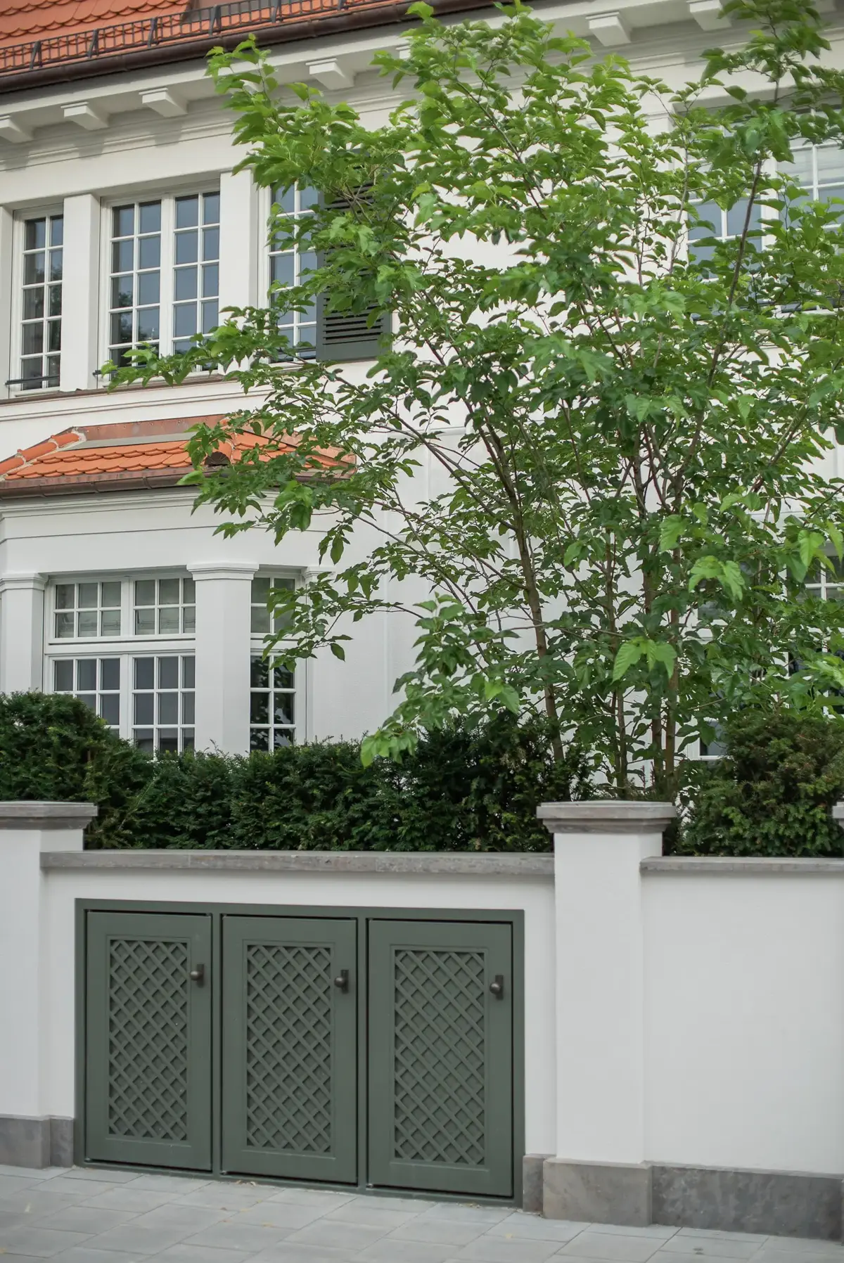 Straßenansicht der Villenfassade mit geschnittener Heckeneinfassung, jungem Blütenbaum und integrierten Mülltonnenboxen in der Gartenmauer.
