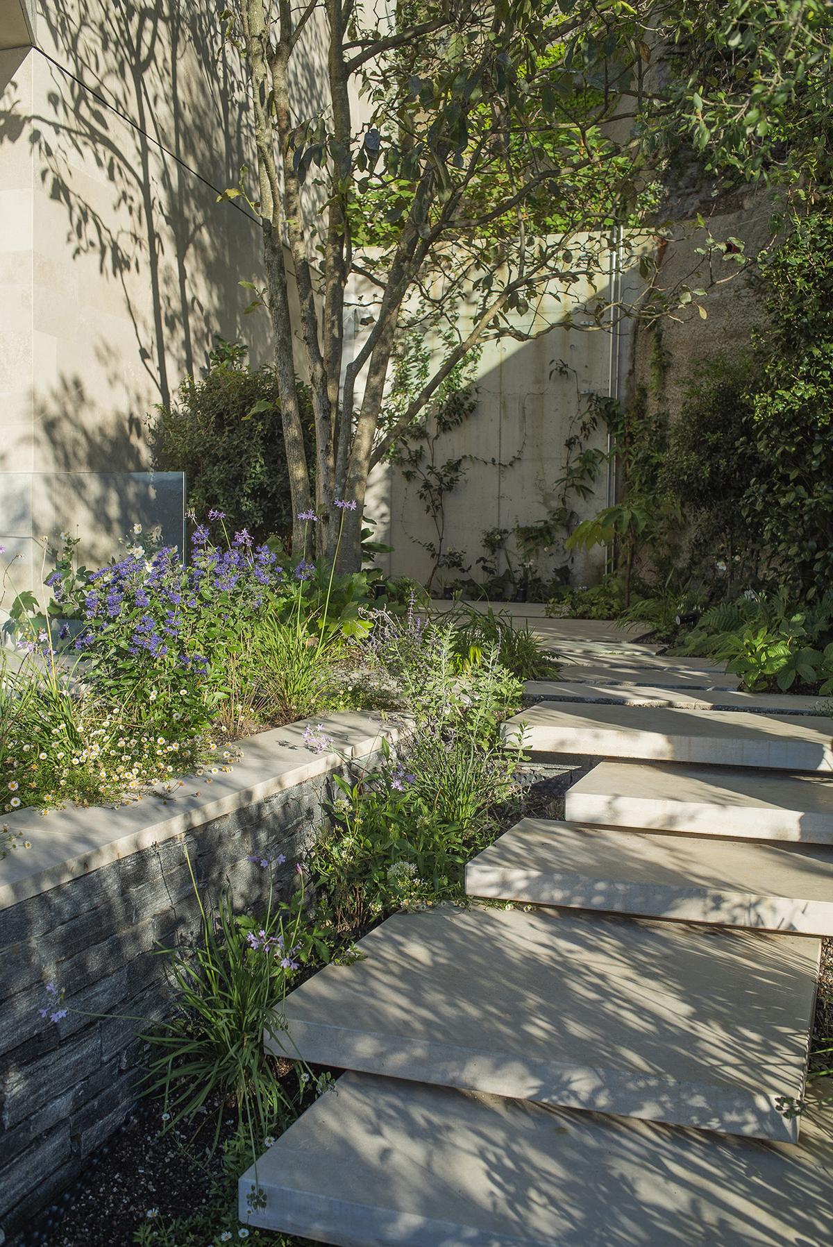 Moderner Garten mit Natursteinplatten, mediterranen Pflanzen und Schattenwurf unter Bäumen.